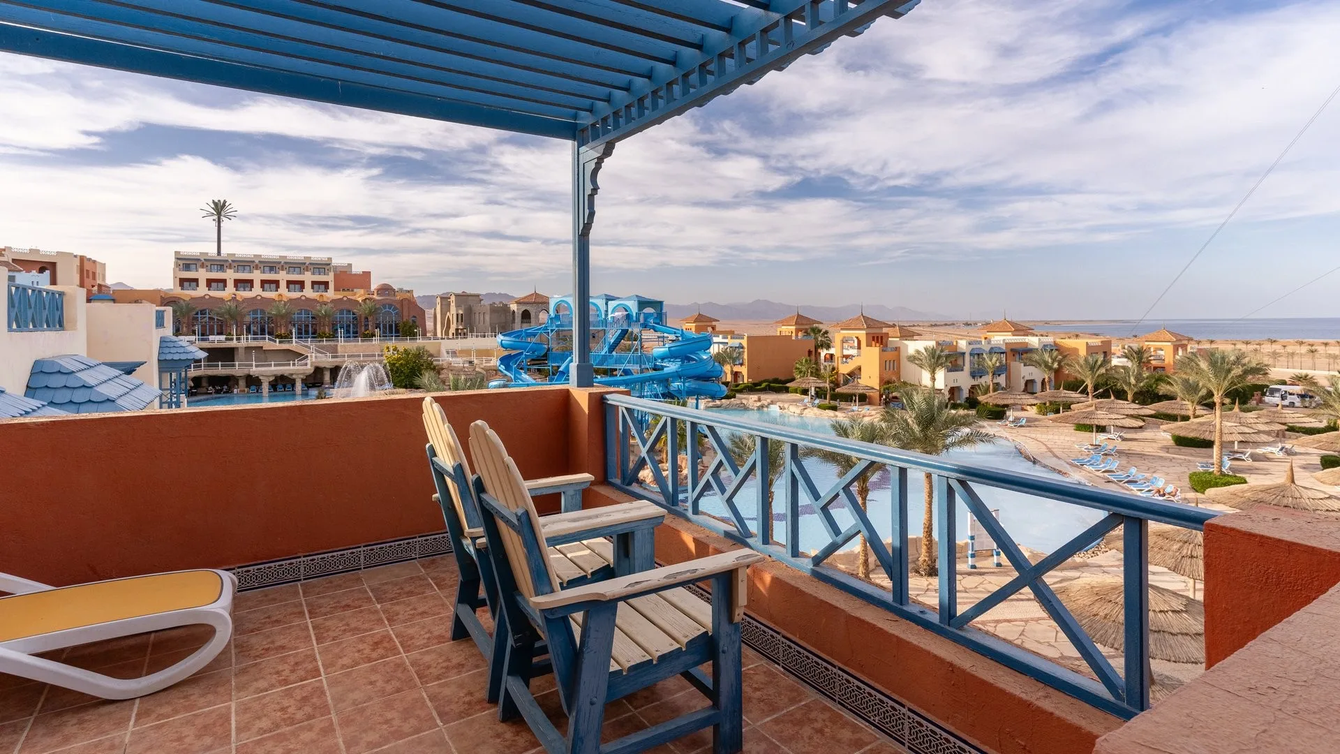 Balcone della camera con sedie in legno e vista sulla piscina con scivoli e sul mare