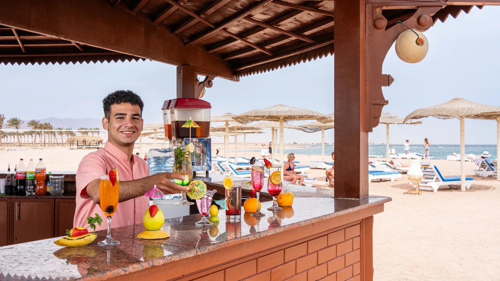Barista sorridente prepara cocktail colorati al beach bar con vista sulla spiaggia e sul mare