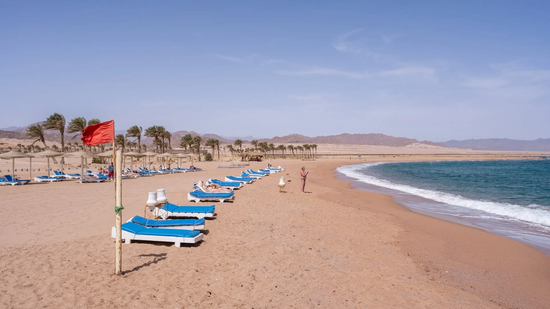 Spiaggia del resort con lettini blu, palme e bandiera rossa, vista sul mare e sulle montagne