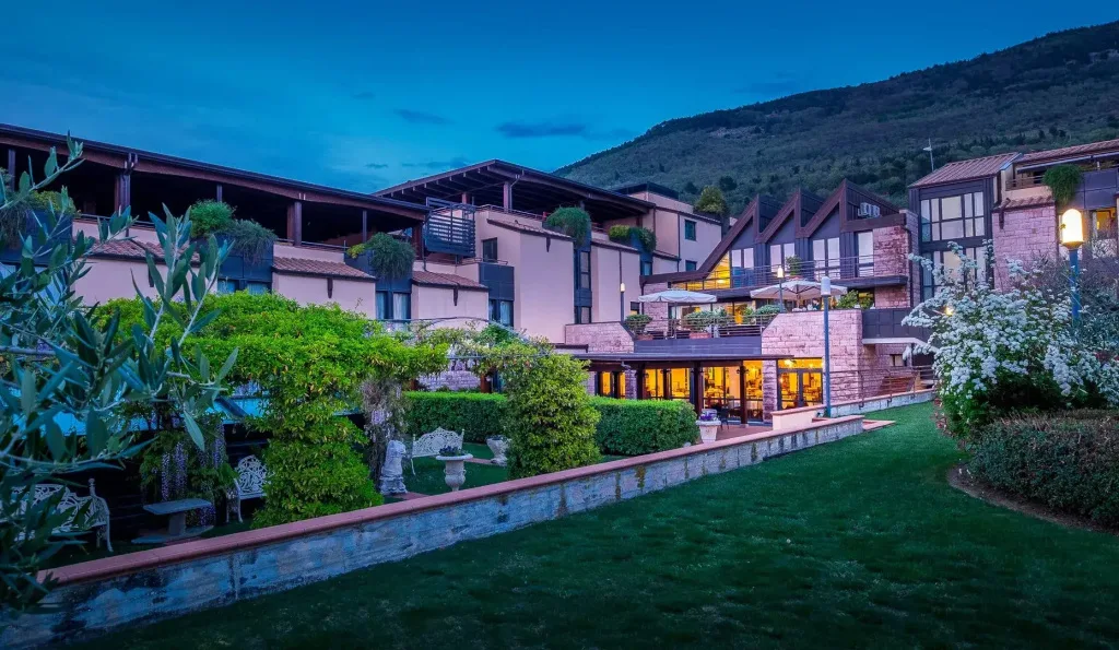 Esterno dell'hotel al crepuscolo con giardino, terrazze illuminate e montagna sullo sfondo