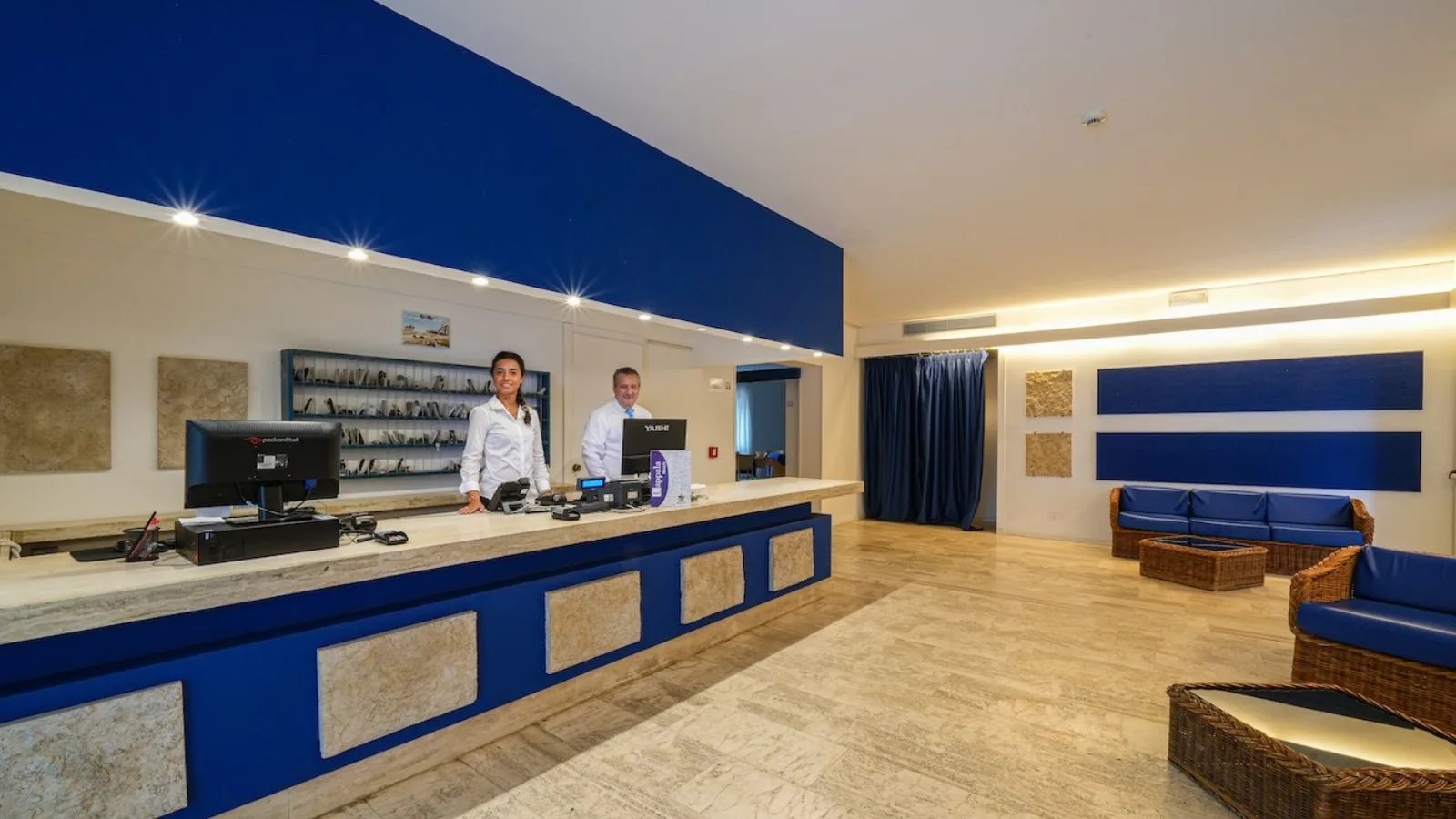 Reception di TH Lacona con banco di accoglienza blu e area salotto nella hall.