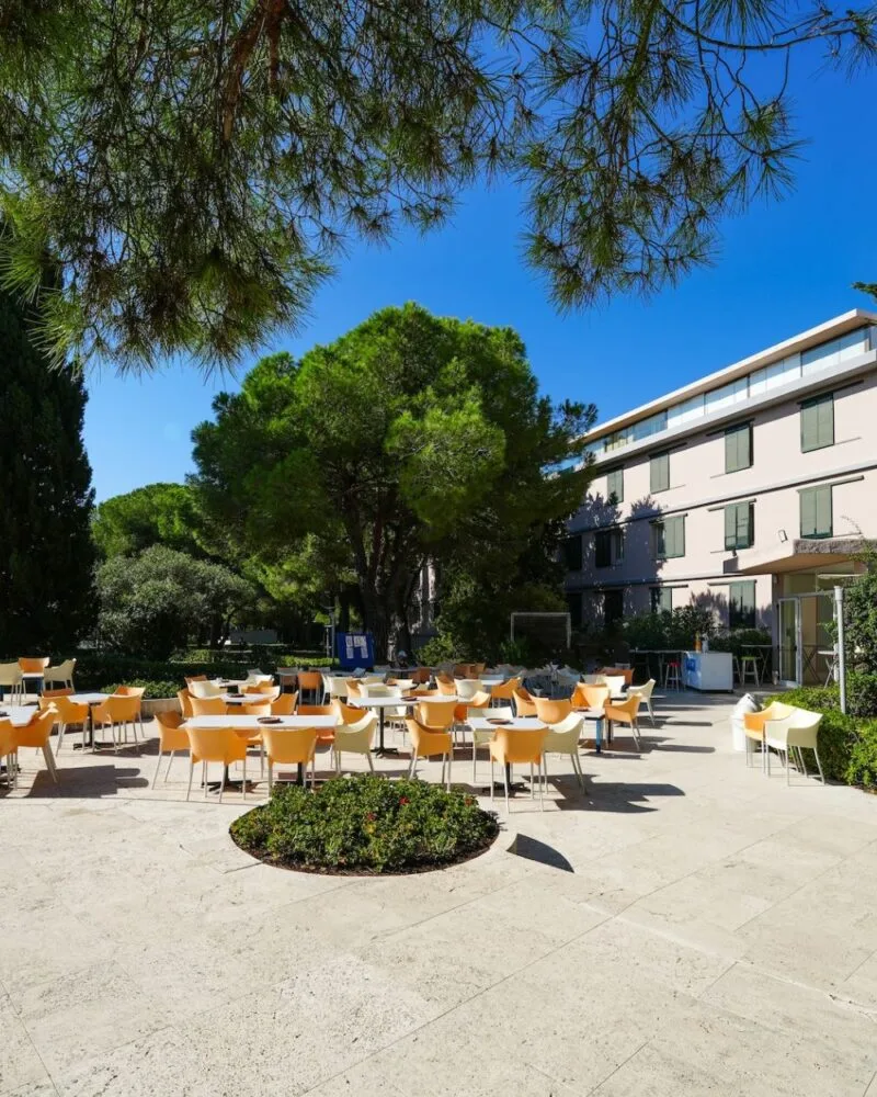 Tavoli e sedie nello spazio esterno di TH Lacona, tra pini e aiuole davanti all’edificio.