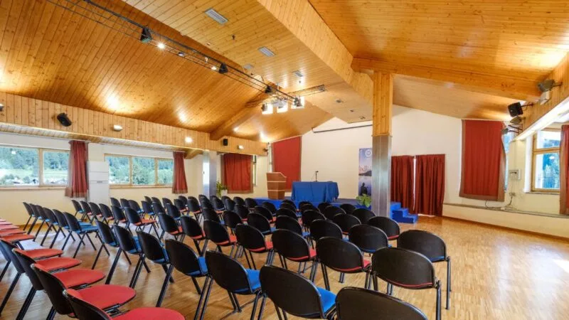 Sala conferenze con sedie nere e rossa, palco con podio e schermo, soffitto in legno e ampie finestre.