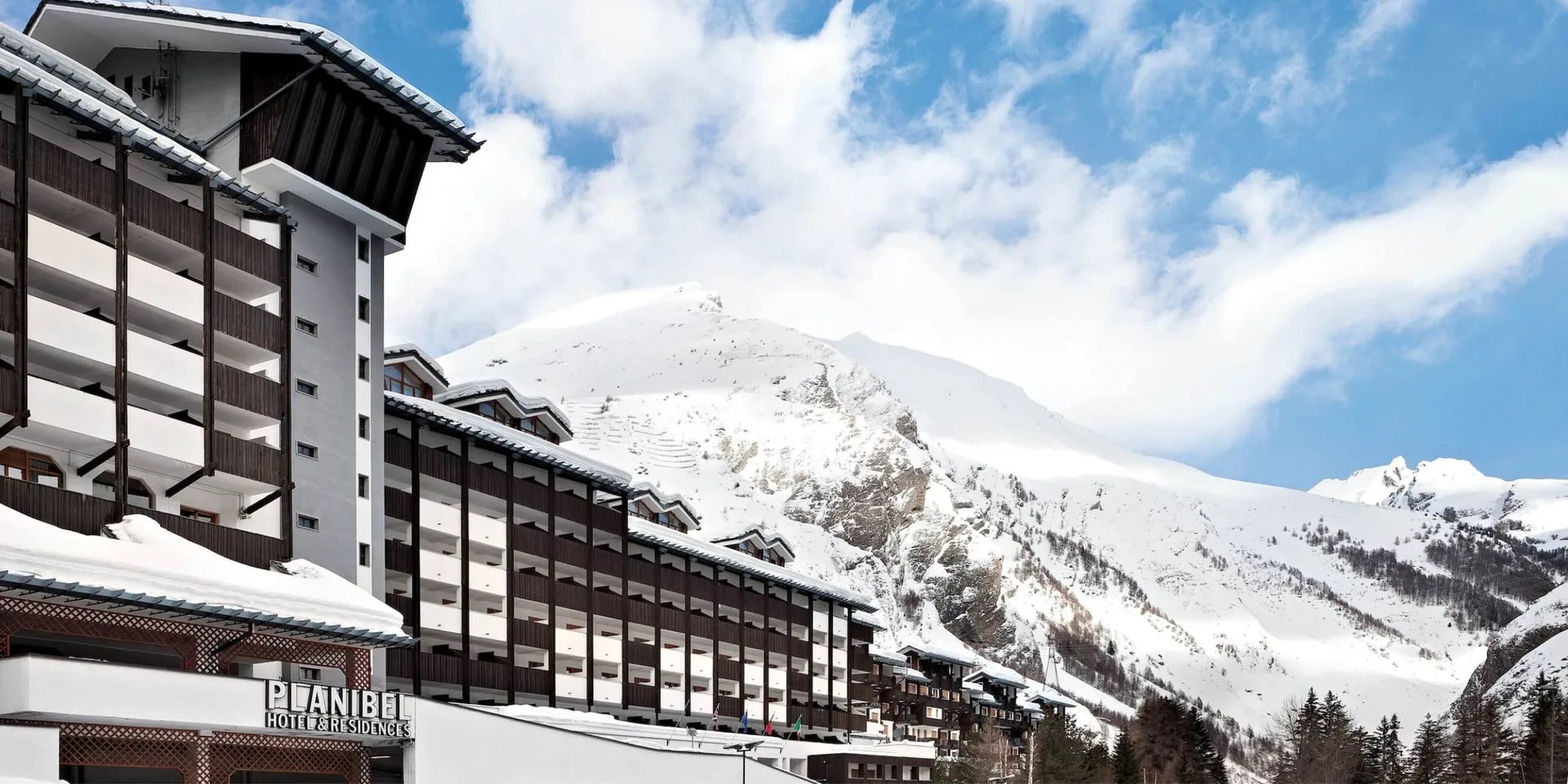 Hotel Planibel con balconi in legno, innevato, ai piedi di montagne innevate sotto un cielo parzialmente nuvoloso.