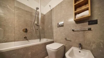 Bagno moderno con vasca, lavabo e wc, pareti rivestite in piastrelle beige e ripiani in legno.