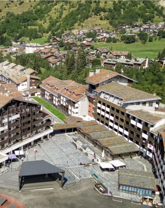 Vista aerea di un complesso di edifici in stile alpino tra gli alberi e le colline verdi.