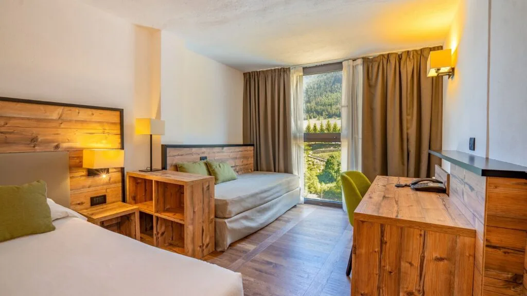 Camera d'albergo luminosa con letti, divano e vista su montagne e foresta attraverso grandi finestr.