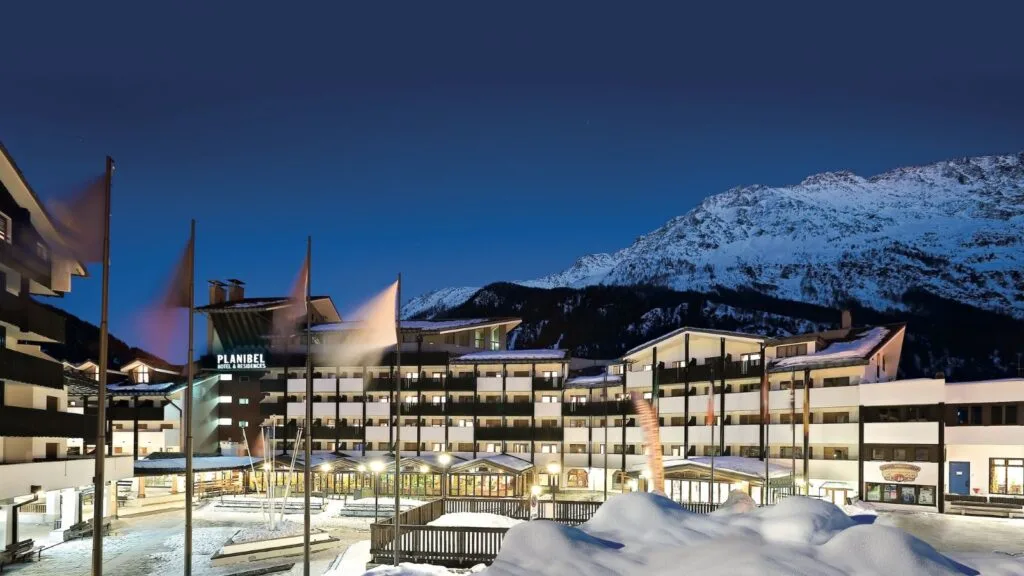 Hotel Resort con montagne innevate e cielo serale, edifici moderni e bandiere nel centro.
