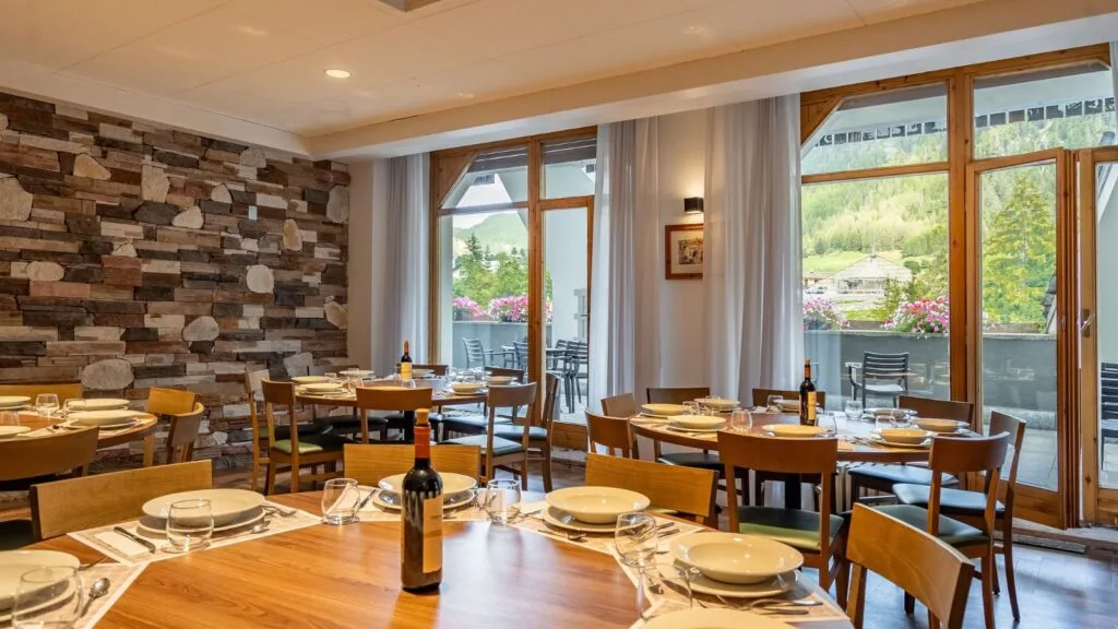 Interno di ristorante con tavoli pronti, parete in pietra e grandi finestre con vista su montagne e fiori.
