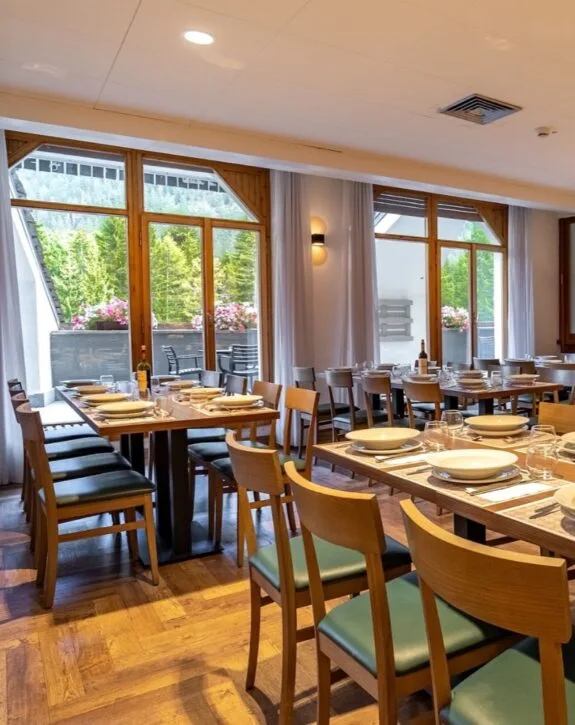 Sala da pranzo con tavoli pronti, grandi finestre e vista sulla natura con fiori e alberi.