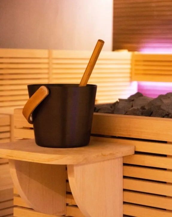 Vaschetta nera per sauna con manici di legno e bastoncino di legno, in ambiente di spa con pannelli di legno.