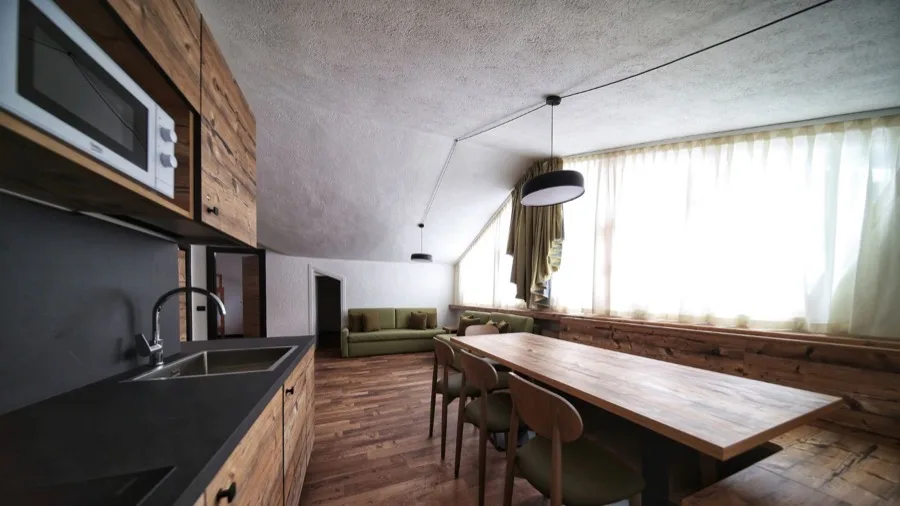 Interno di soggiorno con tavolo in legno, divano e grande finestra con tende chiare.
