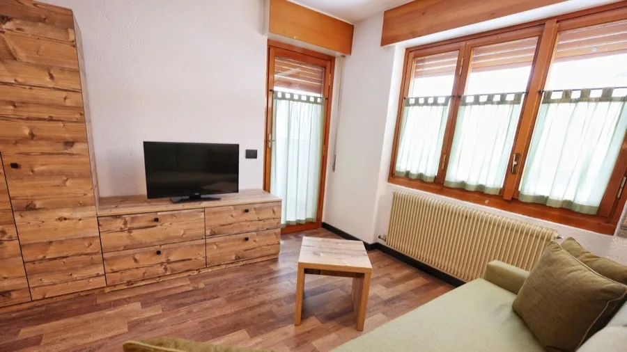 Soggiorno con mobili in legno, televisore, grandi finestre con tende verdi e un divano beige.