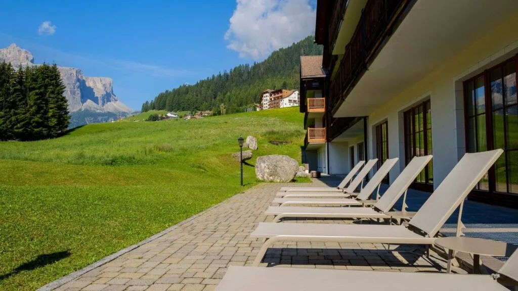 Lettini in fila su patio di hotel con vista su prato verde, montagna e cielo azzurro.