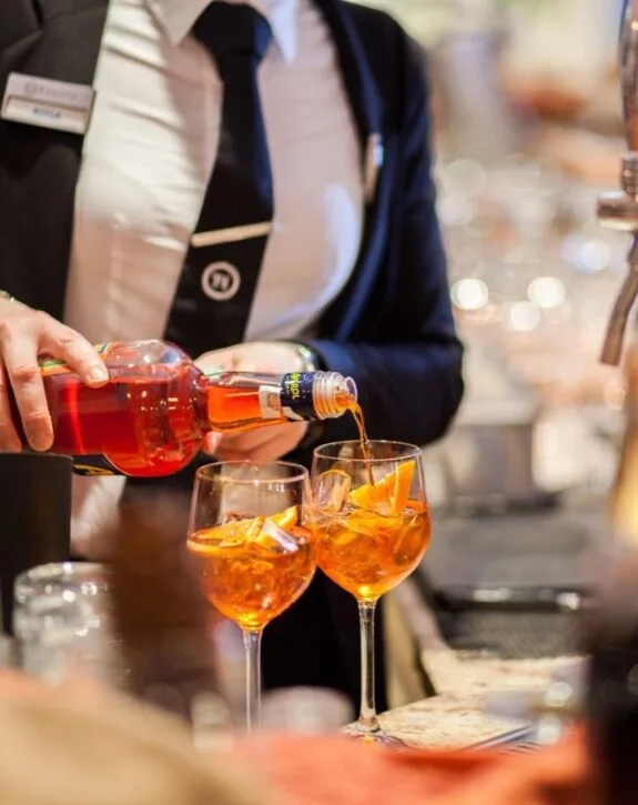 Una persona versa un aperitivo arancione in due bicchieri da vino con ghiaccio e fette di arancia.