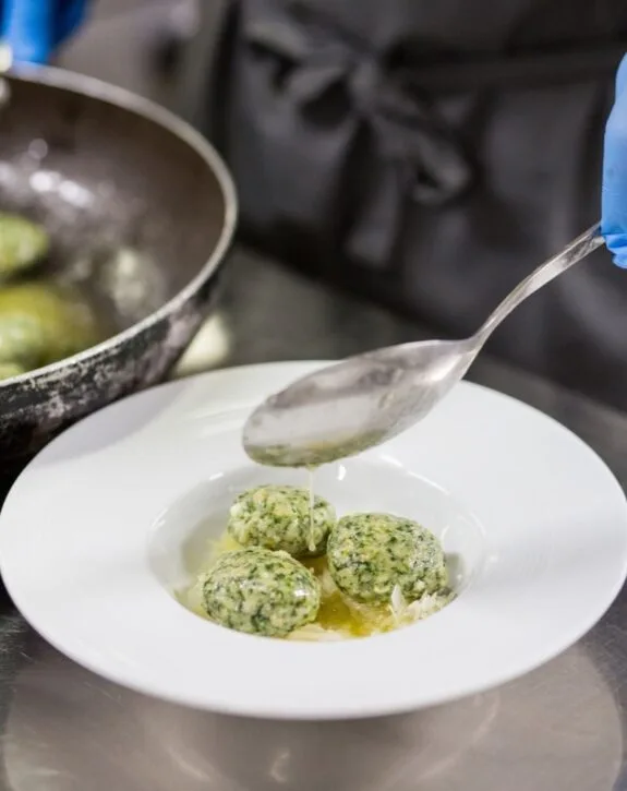 Tre gnocchi verdi conditi con salsa versata da un cucchiaio in un piatto bianco, con padella sullo sfondo.