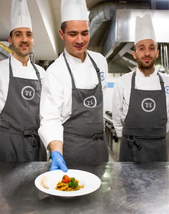 Tre chef con divisa bianca e grembiule grigio presentano un piatto gourmet in cucina professionale.