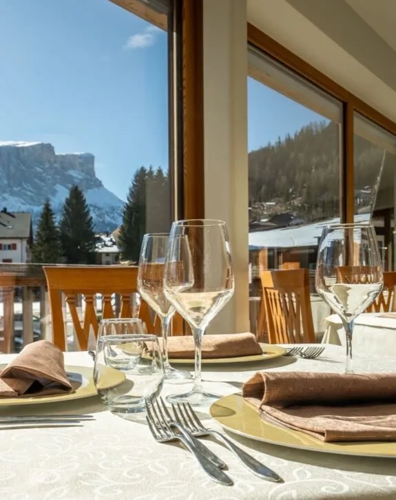 Tavola apparecchiata con bicchieri, posate e tovaglioli davanti a grandi finestre con vista montagna innevata.
