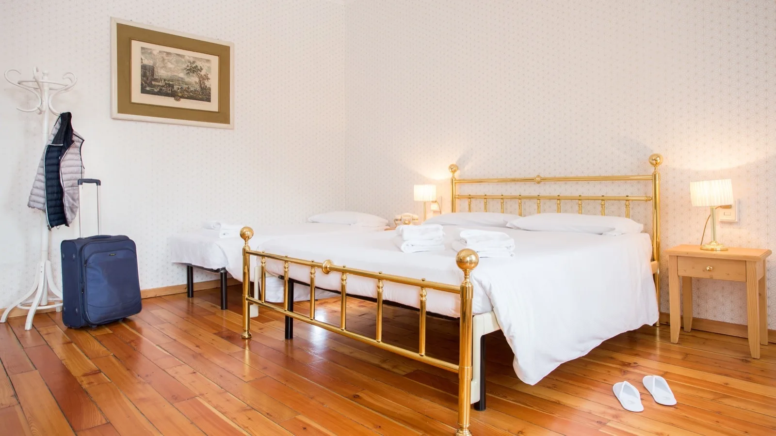 Camera da letto con letto matrimoniale in metallo dorato, pavimento in legno, valigia blu, appendiabiti e pantofole bianche.