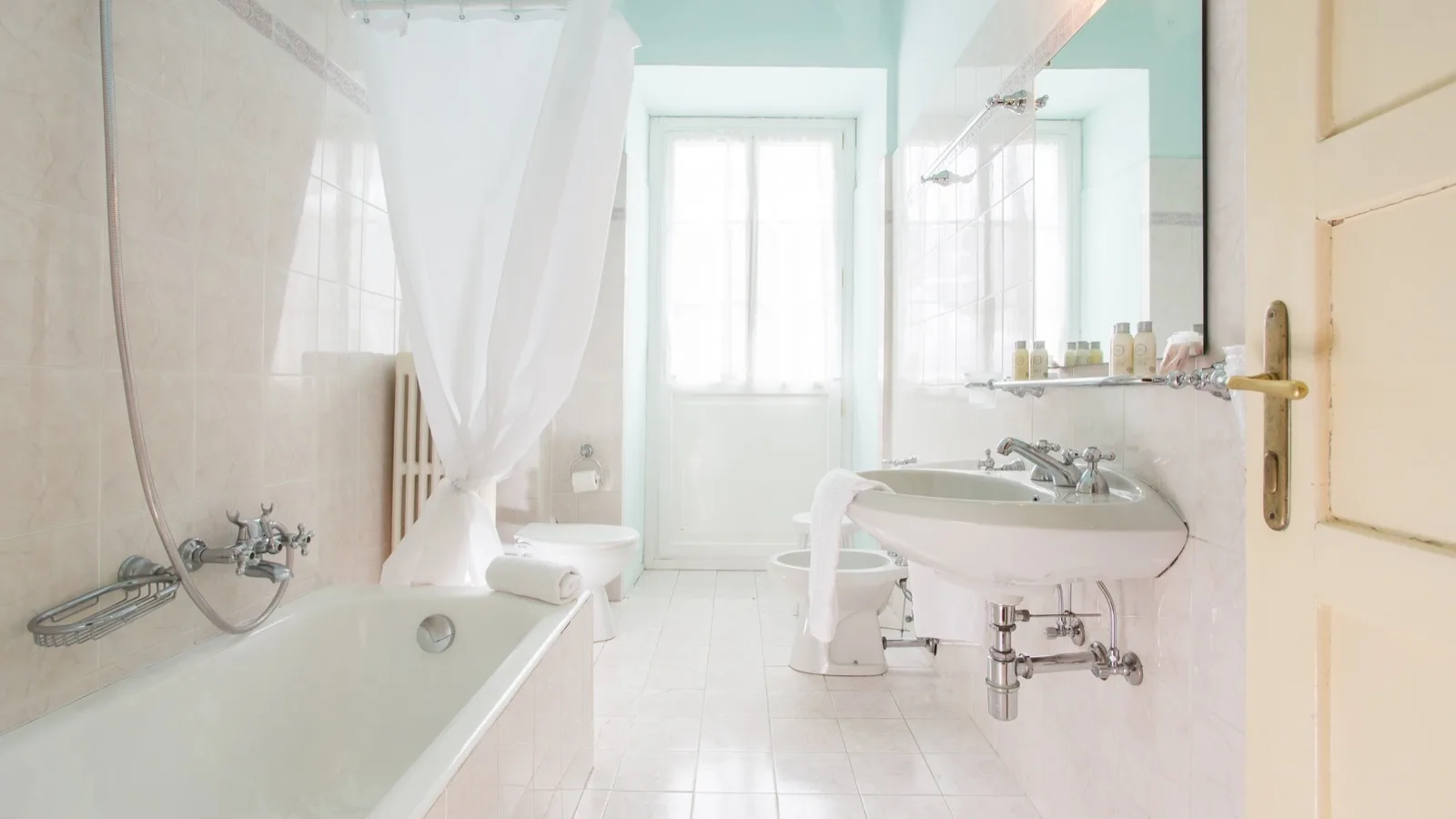 Bagno luminoso con vasca, lavabo, bidet, WC, specchio e accessori bianchi su piastrelle chiare.