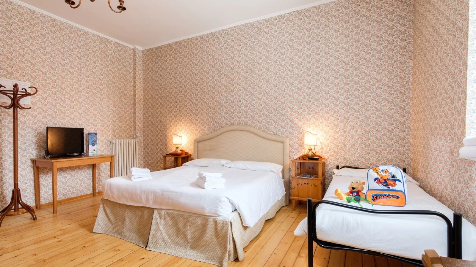 Camera con letto matrimoniale e singolo con zaino e peluche, mobili in legno e pareti con carta da parati floreale.