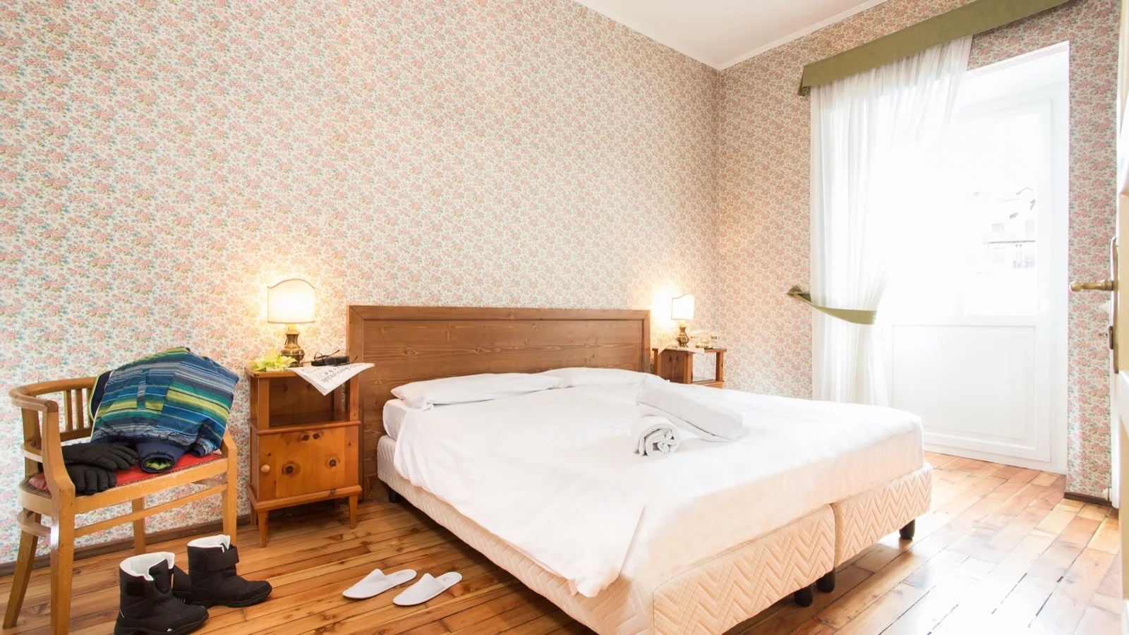 Camera da letto luminosa con letto matrimoniale, carta da parati floreale, pavimento in legno e abbigliamento su sedia.