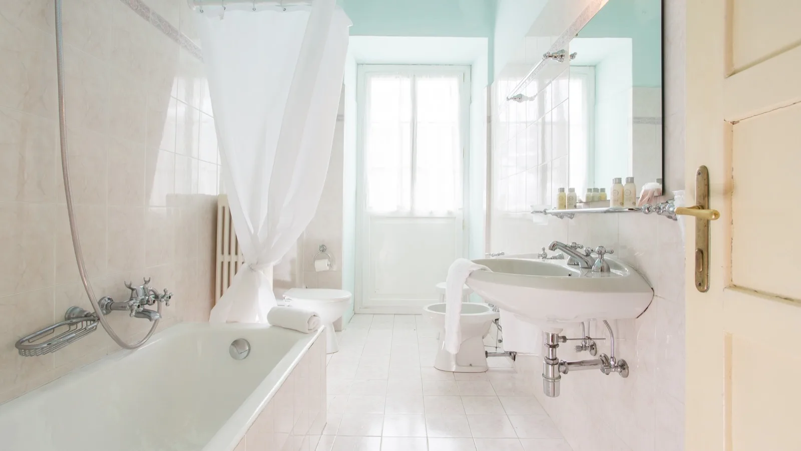 Bagno luminoso con vasca da bagno, lavabo con specchio, water, bidet e finestra con tenda bianca.