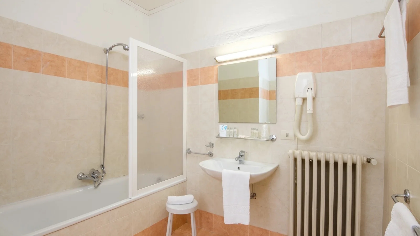 Bagno con vasca, doccia, specchio, phon, lavandino, asciugamani bianchi e piastrelle beige e arancioni.