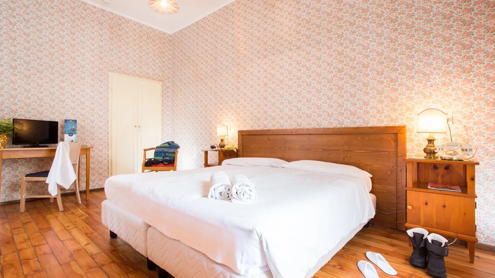 Camera da letto con letto matrimoniale bianco, carta da parati floreale, parquet, scrivania, lampade e stivali neri a terra.
