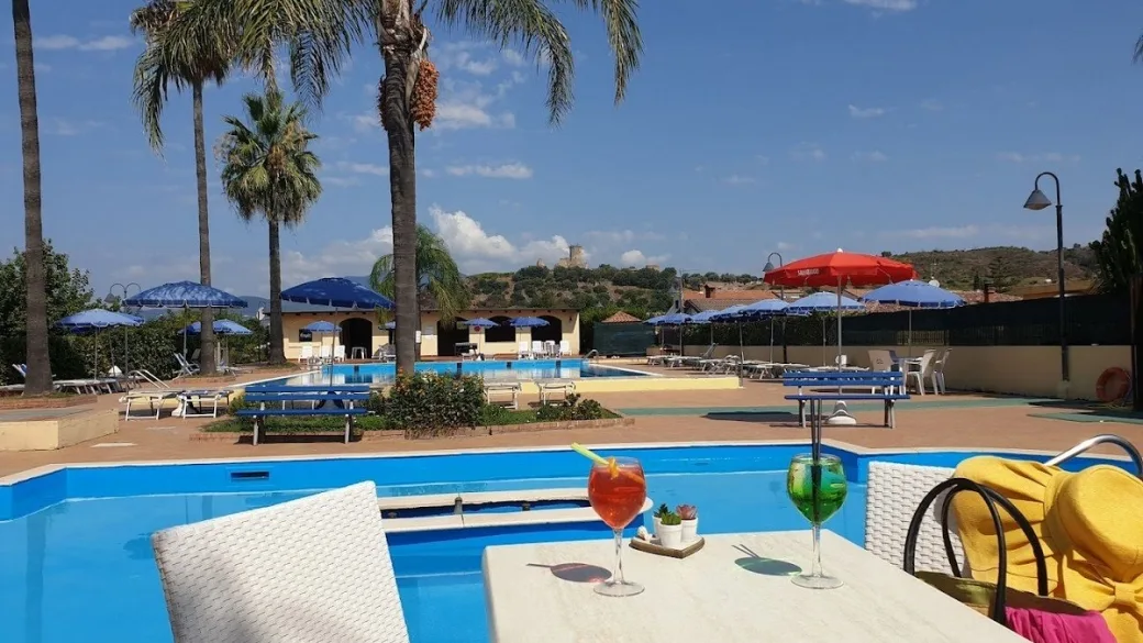 Aperitivo a bordo piscina con cocktail colorati su tavolino e vista su palme e ombrelloni blu