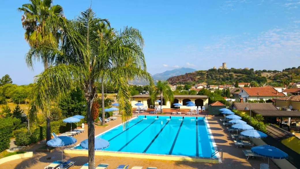 Vista panoramica dall'alto della grande piscina con ombrelloni blu, palme e colline con torre sullo sfondo