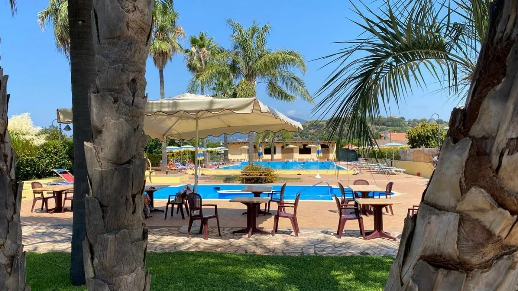 Piscina con ombrellone bianco e tavoli vista tra i tronchi delle palme nel giardino