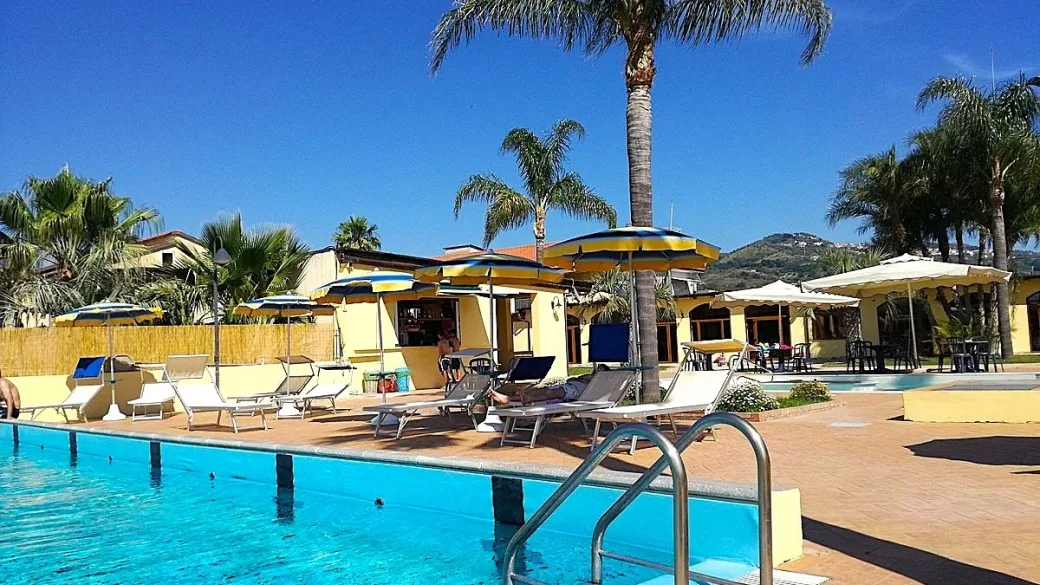Bordo piscina con lettini, ombrelloni gialli e blu, bar e ospiti che prendono il sole