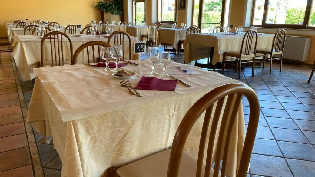 Sala ristorante interna con tavoli apparecchiati con tovaglie chiare, calici e sedie in legno