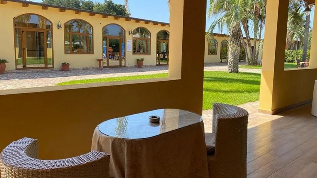 Area lounge coperta con tavolino, poltrone in vimini e vista sul giardino e gli edifici ad archi