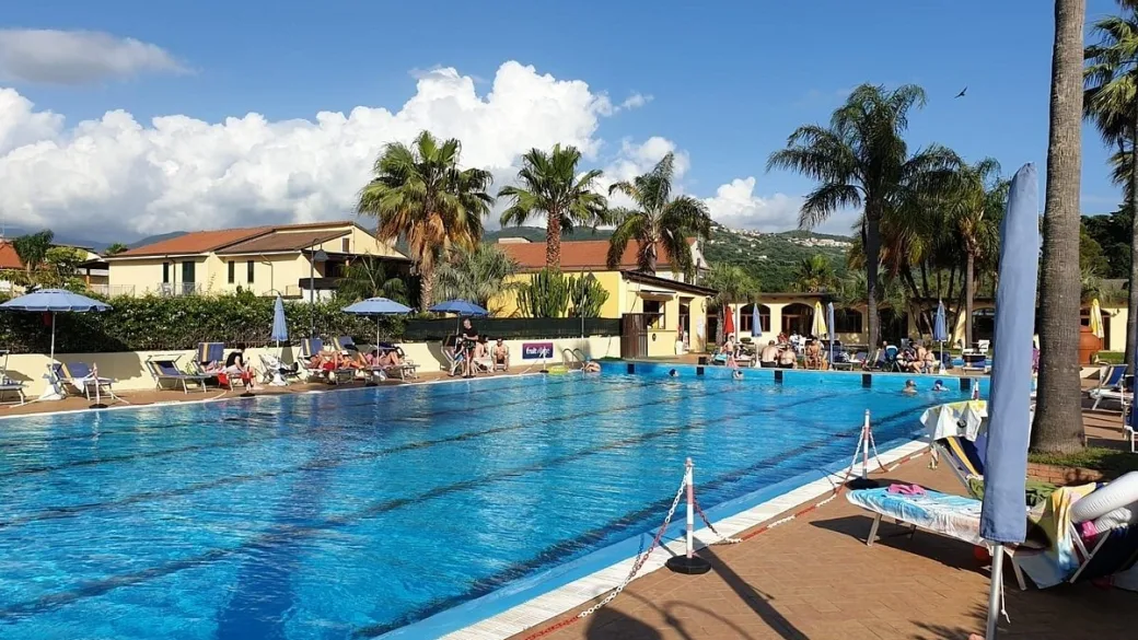 Grande piscina con corsie, ombrelloni blu e numerosi ospiti, circondata da palme ed edifici dell'hotel