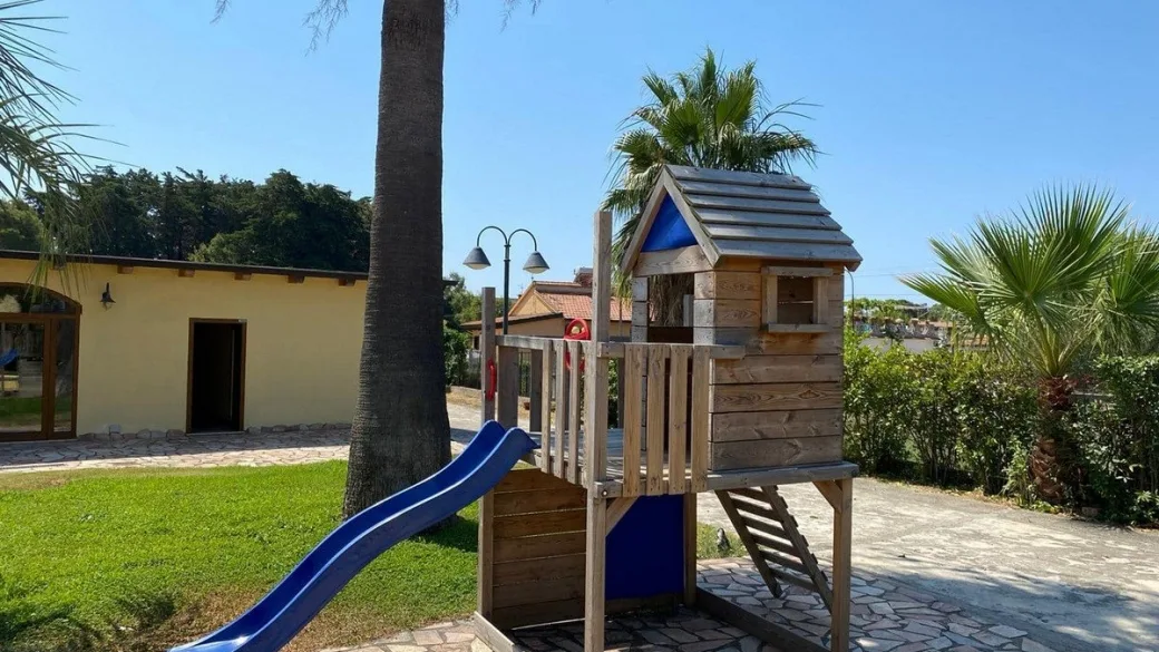Area giochi per bambini con scivolo blu e casetta in legno nel giardino con palme
