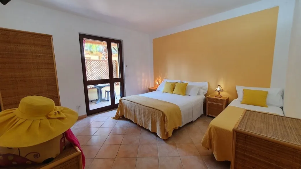 Camera con letto matrimoniale e letto singolo, biancheria bianca e gialla, mobili in vimini e accesso al balcone