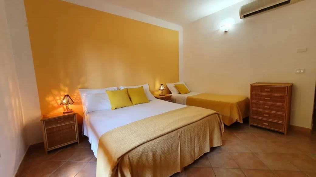 Camera tripla con parete gialla, letto matrimoniale e singolo, comodini con lampade e cassettiera in vimini
