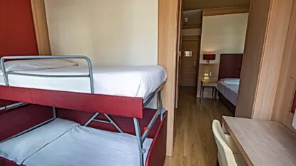 Camera d'albergo con letti a castello rossi, parete bianca, pavimento in legno e scrivania con sedia beige.