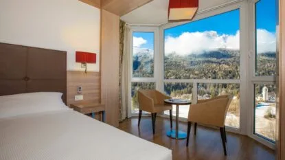 Camera d'hotel con letto matrimoniale e angolo con due poltrone davanti a grandi finestre con vista montagna innevata.