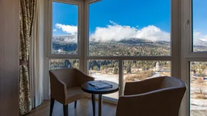 Due poltrone e un tavolino vicino a una finestra con vista su montagne innevate e cielo azzurro.