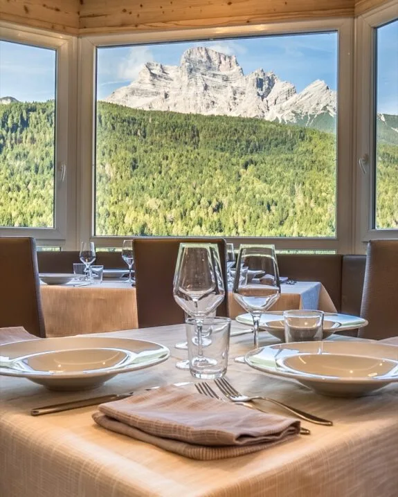 Tavolo apparecchiato in ristorante con vista panoramica su montagne e foresta tramite grandi finestre.