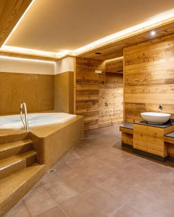 Zona spa interna con vasca idromassaggio rivestita in mosaico marrone, pareti in legno e lavabo moderno su mobile.