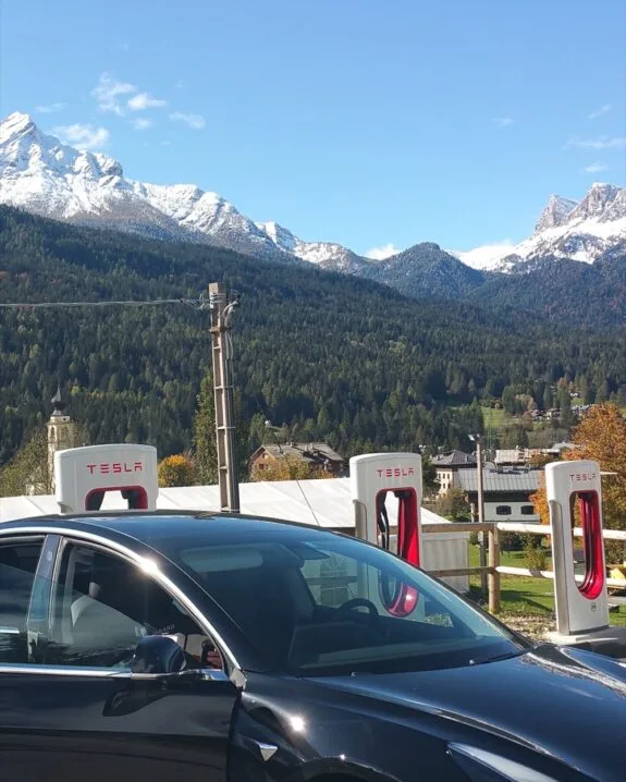 Auto Tesla nera in ricarica davanti a colonnine Tesla con montagne innevate sullo sfondo.