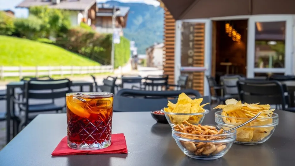 Bicchiere con cocktail, ciotole di snack salati su tavolo all'aperto con sedie e verde sullo sfondo.