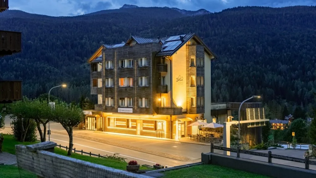 Hotel illuminato di sera ai piedi di una montagna coperta di foresta, con strada e alberi in primo piano.