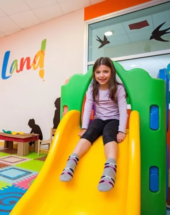 Bambina sorridente con maglietta viola scivola su uno scivolo giallo in area giochi colorata interna.