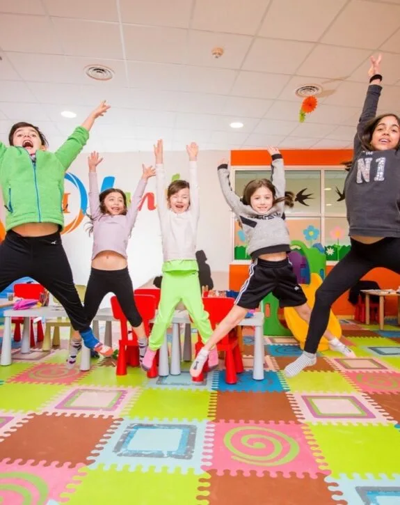 Cinque bambini felici saltano in aria in una stanza colorata con pavimento a tappetini e arredamento per bambini.