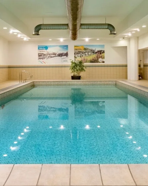 Piscina coperta con piastrelle azzurre, pareti beige, piante e due quadri paesaggistici sul muro.